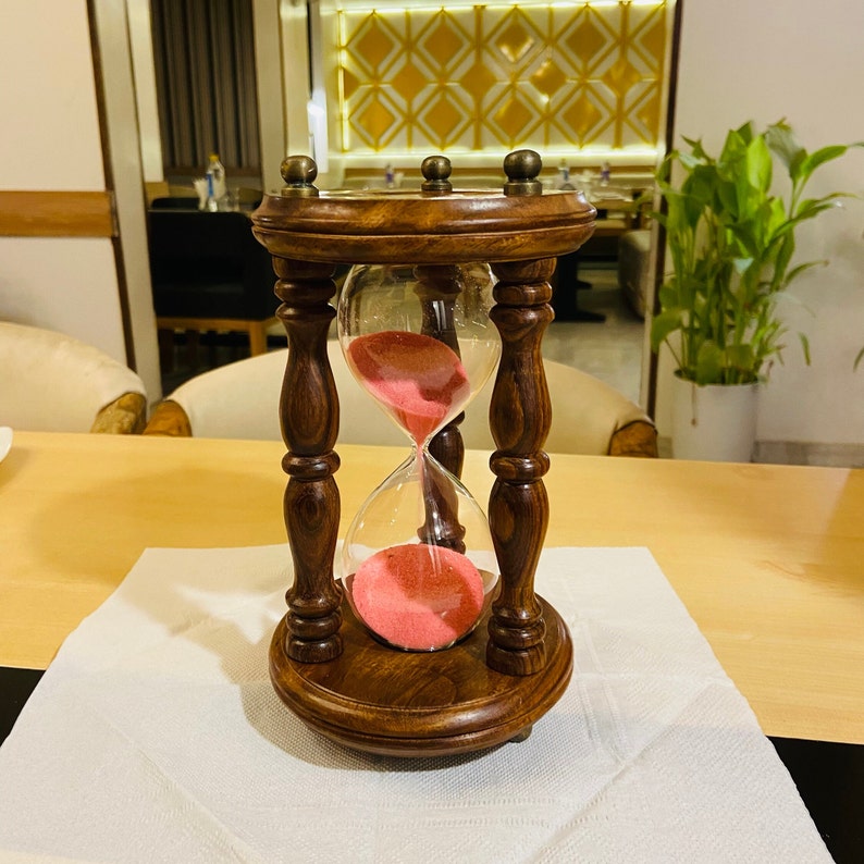 May include: A decorative hourglass with a dark brown wooden frame and base. The glass bulb contains pink sand. The hourglass is set on a white surface, with a light-colored wooden table in the background.
