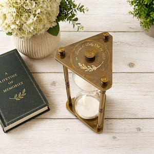 May include: A triangular brass hourglass with white sand, featuring an engraved top plate with the text "Lowin & Billy Oct 14, 2023" and a leaf motif. The frame is supported by three cylindrical pillars, and the glass bulb contains white sand.