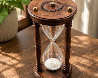 Könnte beinhalten: Eine dunkelbraune Sanduhr aus Holz mit klaren Glaskolben, die weißen Sand enthalten. Der obere runde Holzsockel ist mit drei kleinen messingfarbenen Kugeln und dem eingravierten Text: Rez & Tania, Until the End of Time, sowie einem zentralen Messingknopf versehen.