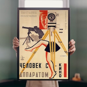 Könnte beinhalten: Ein Vintage-Propagandaposter in Rot, Gelb und Schwarz mit einem Frauenbein und einer Kamera auf einem Stativ. Der Text lautet "Человек с аппаратом кино", was mit "Der Mann mit der Filmkamera" übersetzt werden kann.