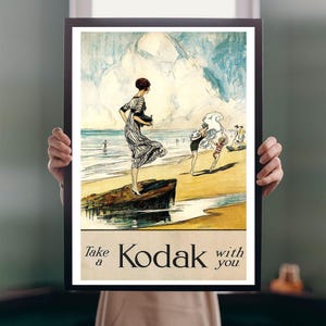 Op de afbeelding: Een ingelijste vintage Kodak-poster. De poster toont een illustratie van mensen op een strand met de tekst "Take a Kodak with you". Het kunstwerk heeft een vintage esthetiek met een kleurenpalet van blauw, geel en bruin. De lijst is zwart.