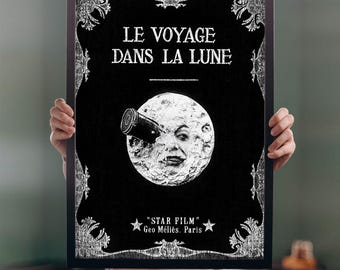 May include: A black and white framed poster featuring the title LE VOYAGE DANS LA LUNE. The center shows a moon with a human face struck by a rocket. Below the moon, the text reads STAR FILM Geo Méliès. Paris, flanked by two white stars.