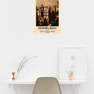 Op de afbeelding: Een vintage reisposter voor Edinburgh, Schotland, met een gedetailleerde illustratie van de architectuur van de stad. De poster heeft een cr&egrave;mekleurige achtergrond met het woord "EDINBURGH" onderaan. De poster wordt op een witte muur weergegeven.