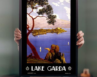 May include: A framed vintage travel poster for Lake Garda, Italy. The poster features a scenic view of the lake with sailboats, mountains, and a castle. The words "LAKE GARDA" are prominently displayed in white text.