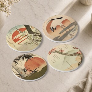 Può includere: Set di quattro sottobicchieri rotondi in ceramica con illustrazioni di arte giapponese. Ogni sottobicchiere presenta una scena diversa con un sole rosso, uccelli e rami nei toni dell'arancione, beige e grigio. Due tazze sono visibili.