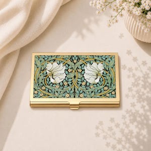 May include: A rectangular metal business card holder with a gold-toned frame and a central panel featuring a repeating botanical pattern of white flowers, green leaves, and small yellow accents against a dark background, with a smooth metallic finish.
