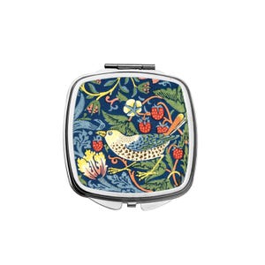 Puede incluir: Un espejo compacto cuadrado, plateado, con apertura de bisagra. La parte superior presenta un diseño estampado colorido de un pájaro, fresas y follaje sobre un fondo azul oscuro. El compacto está cerrado.