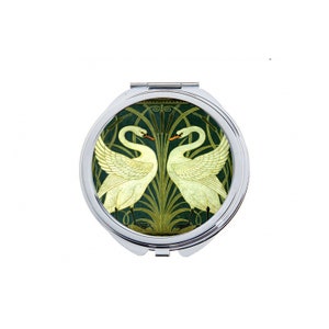 May include: A silver compact mirror with a green and white design featuring two swans facing each other. The swans are surrounded by stylized green reeds.