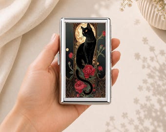 May include: A rectangular silver metal cigarette case featuring a graphic illustration of a black cat sitting in front of a large golden moon, surrounded by red flowers and intricate gold filigree patterns against a dark background.