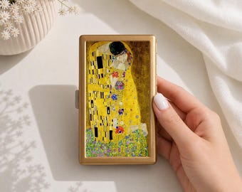 May include: A rectangular gold-toned metal cigarette case featuring a reproduction of Gustav Klimt's The Kiss painting on the front cover. The artwork displays two figures embracing in gold, yellow, and black patterns, set against a floral base.