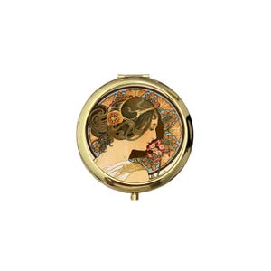 Puede incluir: Un espejo compacto redondo y dorado con una ilustración de estilo Art Nouveau de una mujer con cabello suelto y detalles florales. La imagen está enmarcada por un fondo cálido con tonos anaranjados y marrones.