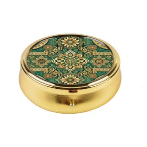 May include: A round, gold-colored pill box with a decorative, ornate design on the lid. The lid features a green and gold floral pattern. The pill box has a small, round knob on the front for opening.