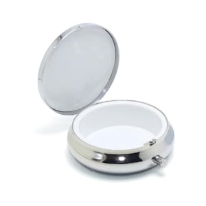 May include: A silver round pill box with a hinged lid. The lid is open and the inside of the box is white.