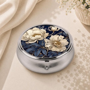Pode incluir: Uma caixa de comprimidos redonda em metal prateado com uma tampa decorada com um fundo azul escuro e flores de peónia em tom creme com detalhes dourados. O recipiente tem um acabamento metálico polido e um pequeno fecho na parte frontal.