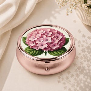 Pode incluir: Uma caixa de comprimidos redonda em metal dourado rosa com uma inserção circular no topo decorada com uma ilustração detalhada de uma flor de hortênsia rosa vibrante com folhas verdes sobre um fundo de cor creme.