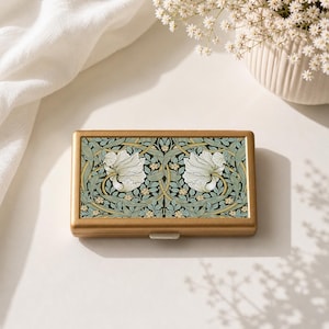 May include: A gold-coloured rectangular case with a floral pattern. The case has a repeating design of white flowers and green leaves on a dark background. It is closed with a small white clasp.