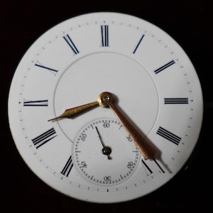 May include: Close-up of a white pocket watch face with black Roman numerals and blue accents. The watch has gold-colored hands and a smaller seconds dial. The hands point to approximately 2:20. The watch face is set against a dark background.