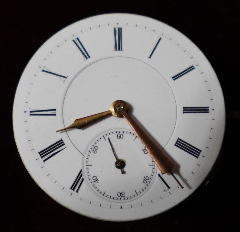 May include: Close-up of a white pocket watch face with black Roman numerals and blue accents. The watch has gold-colored hands and a smaller seconds dial. The hands point to approximately 2:20. The watch face is set against a dark background.