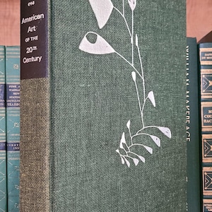 May include: A hardcover book titled "American Art of the 20th Century" by Hunter. The book has a dark green cover with a white floral design and a dark blue spine. The book is standing upright on a wooden shelf.