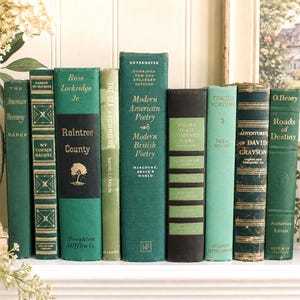 May include: A collection of vintage books with green and gold spines, arranged on a white shelf. Titles include "Raintree County," "Modern American Poetry," and "Roads of Destiny." The books vary in size and style, creating a classic aesthetic.