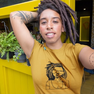 Puede incluir: Una persona lleva una camiseta amarilla mostaza con una ilustración en blanco y negro de una mujer con rastas que lleva gafas de sol y un micrófono. La mujer se muerde el labio y mira hacia un lado. La persona tiene rastas y un aro en la nariz.