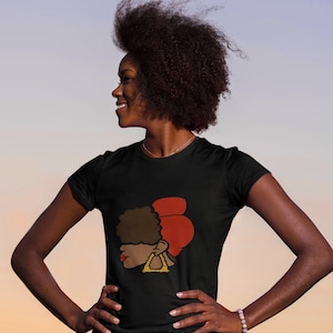 Peut inclure: Un t-shirt noir avec un motif représentant une femme avec un foulard rouge sur la tête et de grandes boucles d'oreilles dorées. La femme regarde vers la droite. Le mannequin porte le t-shirt et a les cheveux noirs bouclés.