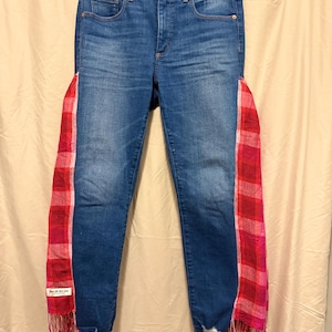May include: Blue denim jeans with red and pink plaid fabric panels down the sides. The jeans have a high waist, a classic five-pocket design, and a frayed hem. The plaid panels have a fringed edge.