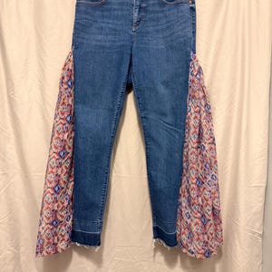 May include: Blue denim jeans with decorative fabric panels on the sides. The panels feature a colorful geometric pattern in shades of pink, blue, and red. The jeans have a classic five-pocket design and a raw hem.