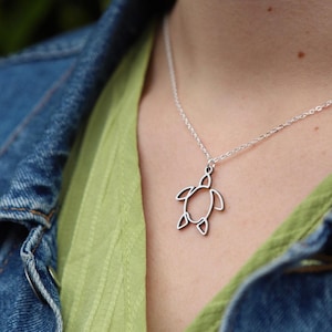 May include: A silver turtle pendant on a necklace. The pendant is a simple outline of a turtle. The necklace is worn over a green shirt and a blue denim jacket.