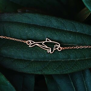 Peut inclure: Un collier délicat en chaîne dorée avec un pendentif en forme de dauphin ajouré. Le collier est présenté sur une feuille vert foncé.