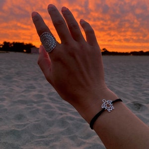 Pode incluir: Um anel de prata com um design circular detalhado e uma pulseira com pingente de tartaruga em um cordão preto. As joias são exibidas em um fundo vibrante de pôr do sol laranja e amarelo.