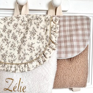 May include: Two small backpacks with a white background and brown accents. One backpack has a floral print and the name "Zélie" embroidered on the front. The other backpack has a gingham pattern.