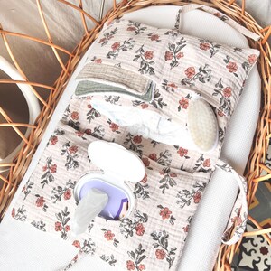 May include: A foldable fabric nappy clutch featuring a floral pattern with muted orange flowers and dark green leaves on a light beige background. The clutch includes a built-in plastic dispenser for wipes and interior pockets holding small cloths.
