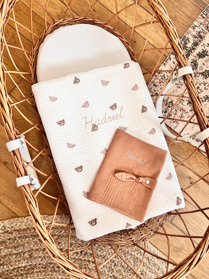 May include: A white woven basket with a white and brown baby blanket with a bear pattern and the name Hadriel embroidered on it. A brown muslin cloth with the name Hadriel embroidered on it is on top of the blanket.