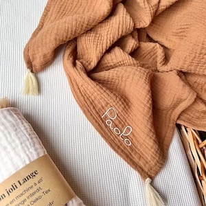 May include: A folded, tan muslin blanket with the name "Paolo" embroidered in white. The blanket has tassels at the corners and is draped over a white and gray striped surface. A rolled white blanket with text is in the foreground.