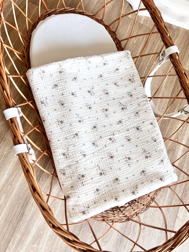 May include: A white wicker bassinet with a white cotton liner and a white and gray floral patterned blanket.