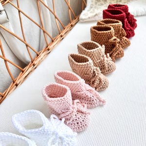 May include: A set of six pairs of crocheted baby booties in various colors, including white, pink, beige, brown, and red. The booties have a lace-up design and are arranged in a row on a white surface.