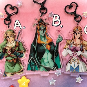 May include: Three acrylic keychains featuring characters from a video game. The keychains are in the shape of the characters and have black heart-shaped clasps. The characters are in various colors, including green, black, and gold. The keychains are labeled A, B, and C.
