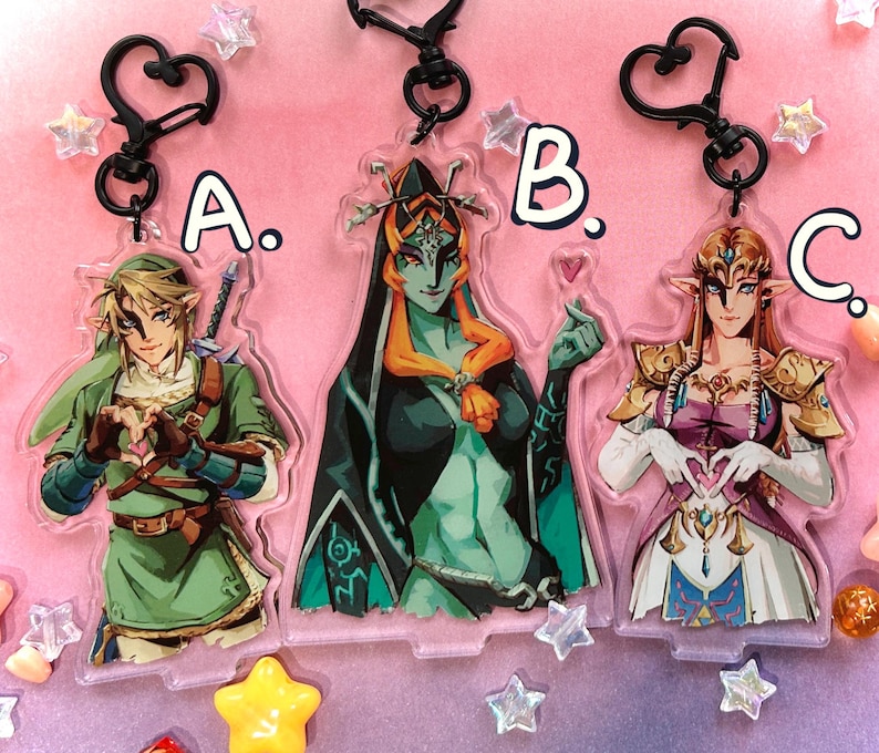 May include: Three acrylic keychains featuring characters from a video game. The keychains are in the shape of the characters and have black heart-shaped clasps. The characters are in various colors, including green, black, and gold. The keychains are labeled A, B, and C.