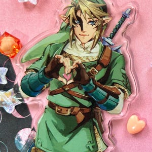 May include: A clear acrylic charm featuring a character from The Legend of Zelda, dressed in green tunic and hat, with a sword on his back. The character is making a heart shape with his hands. The charm has a black clasp.
