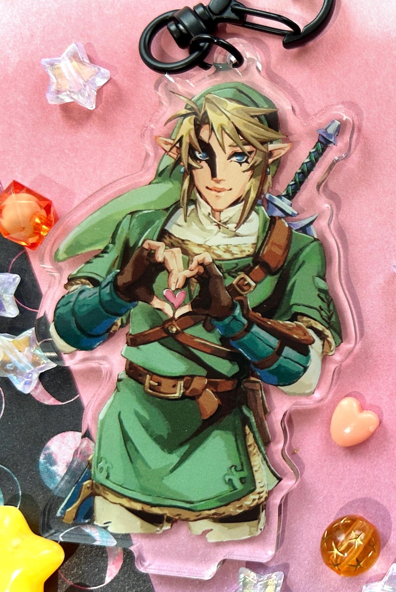 May include: A clear acrylic charm featuring a character from The Legend of Zelda, dressed in green tunic and hat, with a sword on his back. The character is making a heart shape with his hands. The charm has a black clasp.