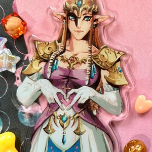 May include: A clear acrylic keychain featuring a character from The Legend of Zelda. The character is depicted with long blonde hair, a purple dress, and gold armor. The character is making a heart shape with their hands. The keychain has a black clasp.