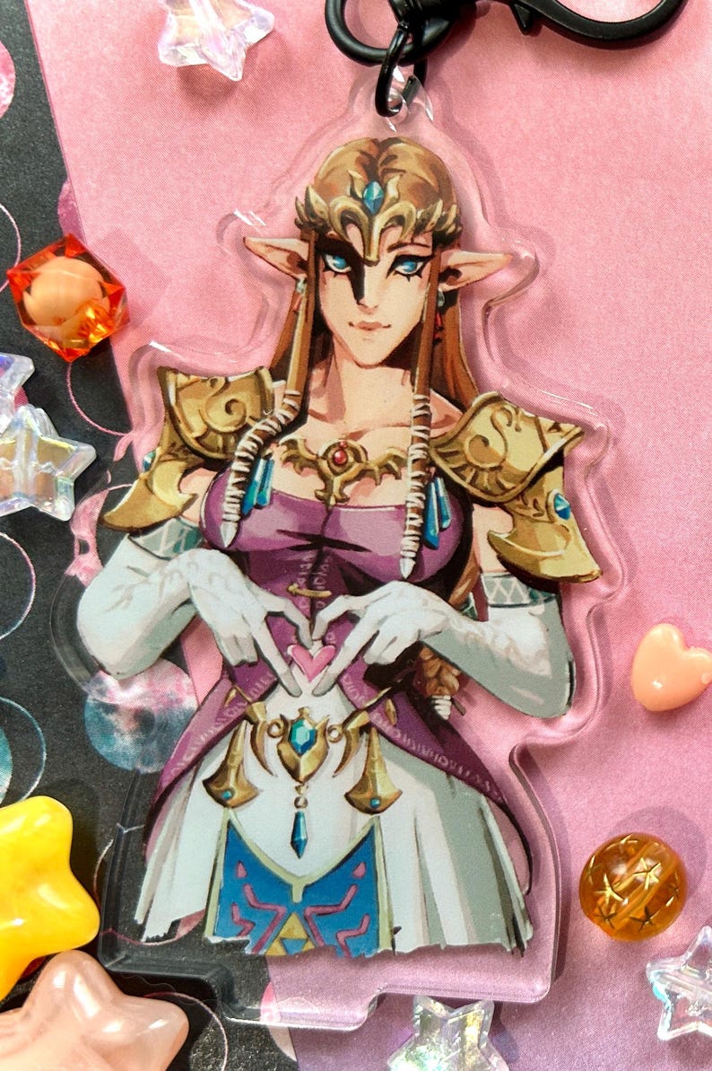 May include: A clear acrylic keychain featuring a character from The Legend of Zelda. The character is depicted with long blonde hair, a purple dress, and gold armor. The character is making a heart shape with their hands. The keychain has a black clasp.