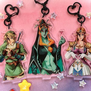 May include: Three acrylic keychains featuring characters from The Legend of Zelda. The keychains depict Link in green, Midna in black and orange, and Zelda in white and purple. Each keychain has a black heart-shaped clasp and is surrounded by star-shaped decorations.