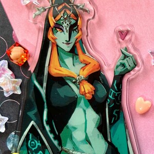 May include: A clear acrylic charm featuring a teal-skinned character with orange hair and a black outfit. The character is adorned with intricate details and a stern expression. The charm is decorated with a heart and star-shaped embellishments.