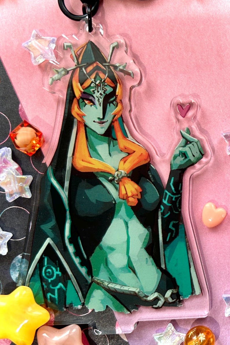 May include: A clear acrylic charm featuring a teal-skinned character with orange hair and a black outfit. The character is adorned with intricate details and a stern expression. The charm is decorated with a heart and star-shaped embellishments.