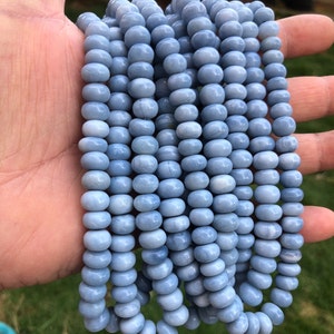 Puede incluir: Hebras de cuentas ovaladas de color azul claro. Las cuentas están ensartadas, creando una apariencia fluida y texturizada. Las cuentas son de un color azul suave y apagado. La imagen está tomada al aire libre.