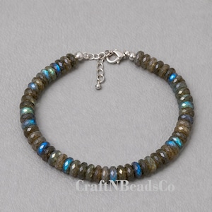 Könnte beinhalten: Ein Armband mit facettierten Labradorit-Perlen in Grau-, Braun- und Blautönen. Das Armband hat einen silberfarbenen Verschluss und ist auf einem flexiblen Band aufgezogen. Das CraftNBeadsCo-Logo ist sichtbar.