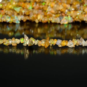 Puede incluir: Una hebra de pequeñas virutas de ópalo irregulares e iridiscentes en tonos dorados, anaranjados y blancos. Las virutas están dispuestas en línea sobre una superficie negra, con sus reflejos visibles debajo.