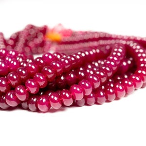 Peut inclure: Gros plan sur un fil de perles de pierres précieuses polies, d'un rouge profond. Les perles sont rondes et de taille uniforme, avec une finition brillante. Le fond est blanc, ce qui met en valeur la couleur et la texture riches des perles. Les perles sont enfilées, suggérant un collier ou un bracelet.